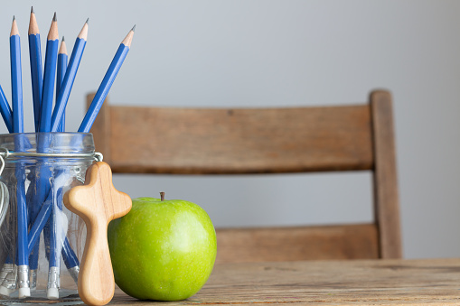 weathered vintage desk with small wood comfort cross, jar of blue pencils and one green apple sitting on the desk top, white background with copy space