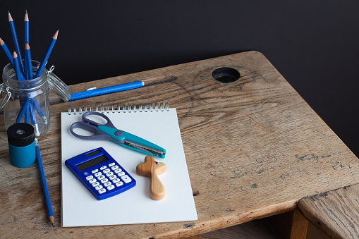 Vintage rustic wood school desk with small christian wood cross and office or school supplies including open notebook, blue calculator, pencils, scissors and pencil sharpener. With black background and copy space.