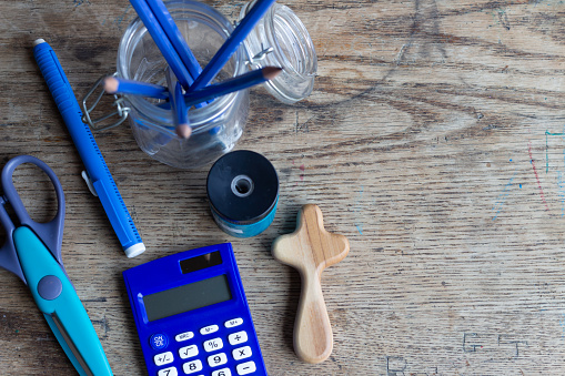 small christian wood cross sitting on desk top of vintage wood desk with blue school supplies including pencils, eraser, calculator, scissors and pencil sharpener with copy space shot from above