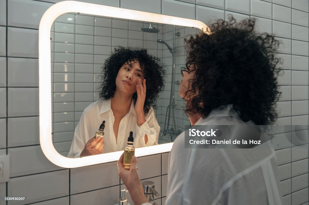 Young mixed race female gently applying face oil serum with fingers in modern bathroom. Young mixed race female gently applying face oil serum with fingers in modern bathroom. Women skincare and beauty concept. Skin Care Stock Photo