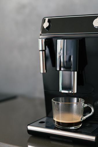 Glass with espresso ready and waiting after coffee making machine finished, hot beverage with foam, black home appliance with metallic parts, clean furniture, blurred background. Vertical shot