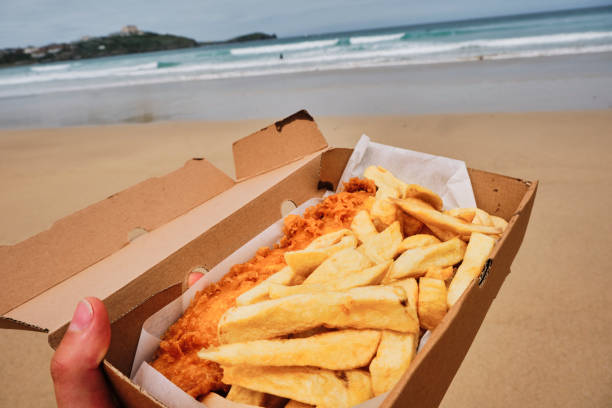 POV of a hand holding a cardboard box filled with traditional Fish and Chips on Towan Beach, Newquay, Cornwall on a June day.