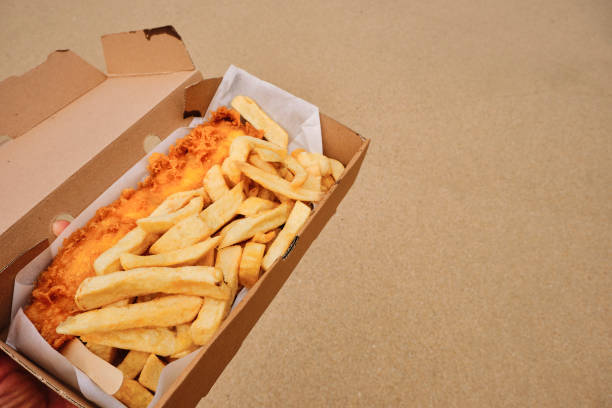 POV of a hand holding a cardboard box filled with traditional Fish and Chips on Towan Beach, Newquay, Cornwall on a June day.