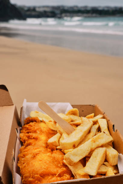 POV of a hand holding a cardboard box filled with traditional Fish and Chips on Towan Beach, Newquay, Cornwall on a June day.