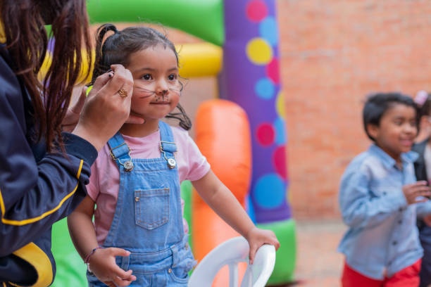 niña de 2 a 3 años sigue mientras la asistente de la fiesta se pinta la cara durante el evento y se prepara para disfrutar y divertirse - 2 3 years children only group of people enjoyment fotografías e imágenes de stock