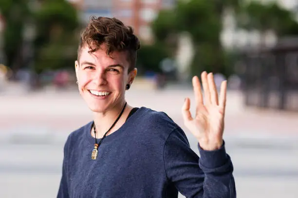 Photo of Non binary transgender Tomboy looking at camera, greeting and waving hand smiling during internet online communication.