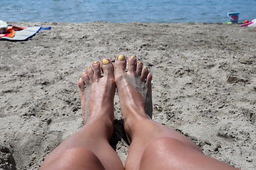 Female legs with yellow pedicure on feet. Women lying in sea sand on the beach on a summer day. Close up, copy space
