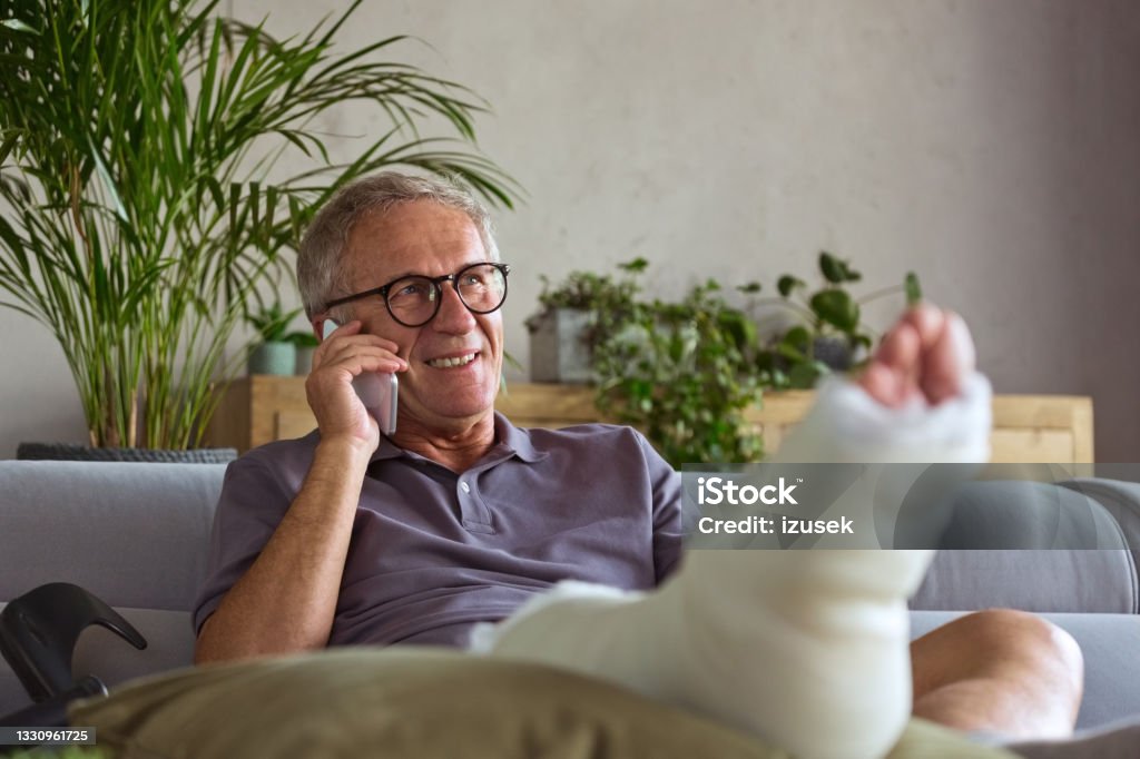 Senior man with broken leg at home Cheerful senior man with broken leg in plaster cast sitting on sofa at home and talking on smart phone. Telephone Stock Photo