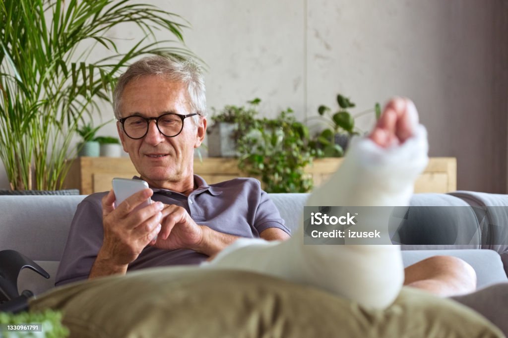 Senior man with broken leg at home Cheerful senior man with broken leg in plaster cast sitting on sofa at home and using smart phone. Physical Injury Stock Photo