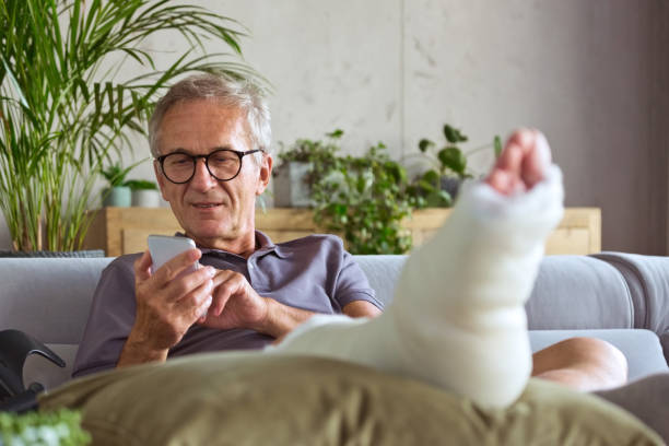 Senior man with broken leg at home Cheerful senior man with broken leg in plaster cast sitting on sofa at home and using smart phone. orthopedic cast stock pictures, royalty-free photos & images