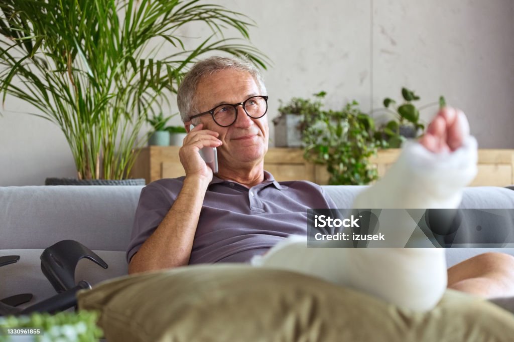 Senior man with broken leg at home Cheerful senior man with broken leg in plaster cast sitting on sofa at home and talking on smart phone. Orthopedic Cast Stock Photo