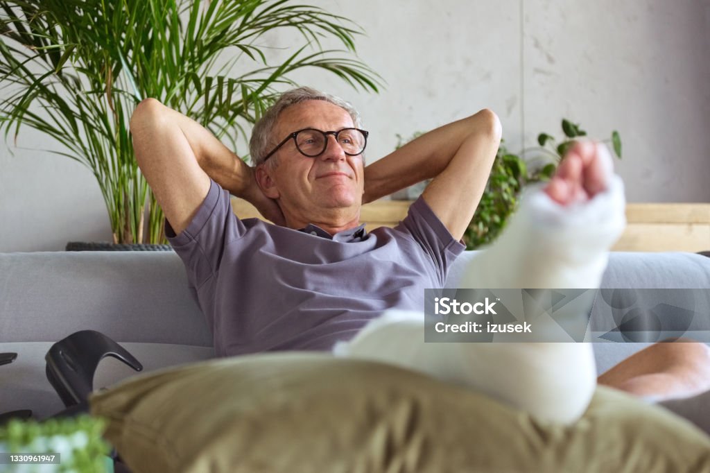Senior man with broken leg at home Cheerful senior man with broken leg in plaster cast sitting on sofa at home, resting with raised arms. Orthopedic Cast Stock Photo