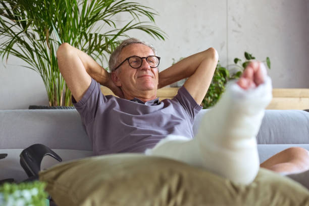 Senior man with broken leg at home Cheerful senior man with broken leg in plaster cast sitting on sofa at home, resting with raised arms. broken leg stock pictures, royalty-free photos & images