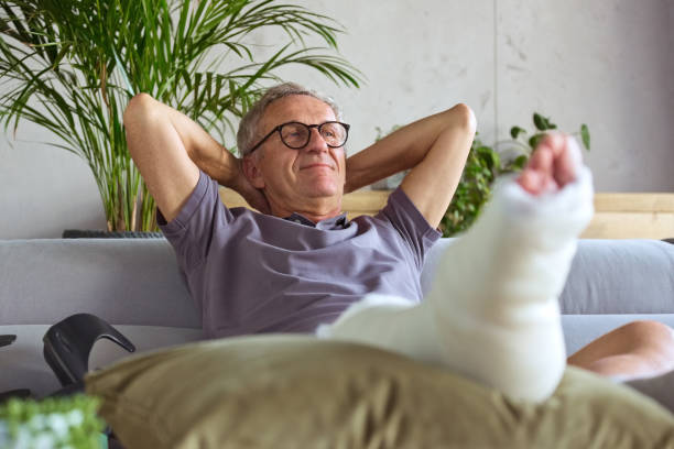 Senior man with broken leg at home Cheerful senior man with broken leg in plaster cast sitting on sofa at home, resting with raised arms. man sitting with a bandage on his leg stock pictures, royalty-free photos & images