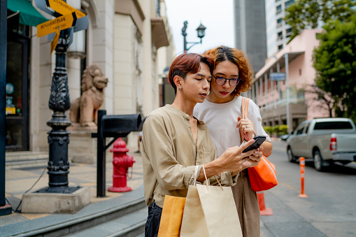 Asian LGBTQIA couple in casual clothing waiting for the car after shopping in downtown city.