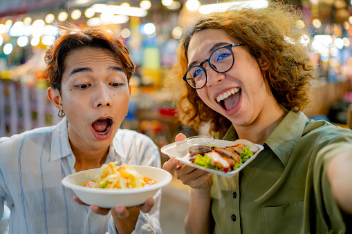 Asian LQBTQIA couple using smart phone taking a selfie with Thai food at night market.