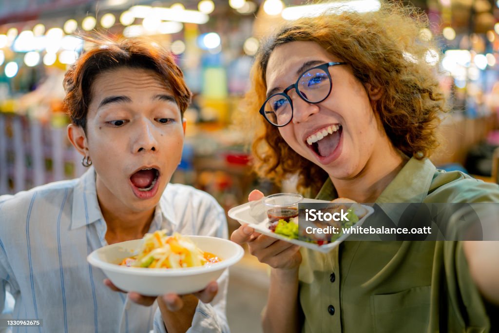 Two vlogger filming a video with their street food. Asian LQBTQIA couple using smart phone taking a selfie with Thai food at night market. Street Food Stock Photo