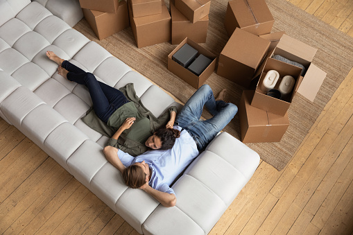 First day at new home. Above view of young spouses happy owners renters of modern apartment relax talk share plans ideas. Affectionate family couple cuddle on couch among carton boxes dream together
