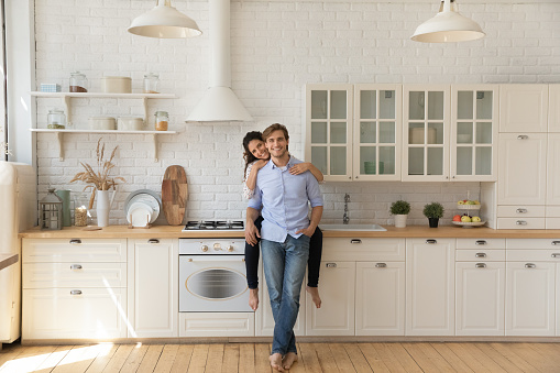 Portrait of affectionate young wife sitting on kitchen counter hug smiling husband from behind at modern renovated interior. Loving young couple homeowners look at camera glad of buying new furniture