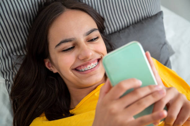 ベッドに横たわっている彼女のスマートフォンを使用してブレースを持つ笑顔の十代の女の子の肖像画 - number 13 写真 ストックフォトと画像