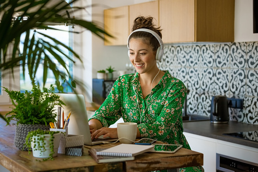 Cheerful young beautiful woman wearing green dress and headphone sitting at table in the kitchen and working at home, having video conference.