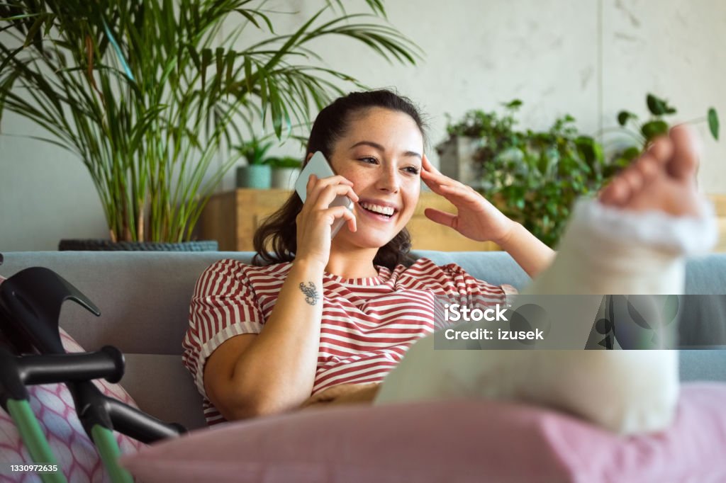 Cheerful young woman with broken leg on phone Smiling young man with broken leg in plaster cast lying down on sofa at home and talking on smart phone. Physical Injury Stock Photo