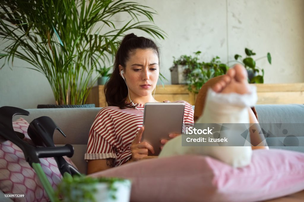 Displeased young woman with broken leg at home Disappointed young man with broken leg in plaster cast lying down on sofa at home and using a digital tablet, having video call. 20-24 Years Stock Photo