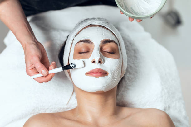 cosmetologist applying the alginates facial mask to woman while lying on a stretcher in the spa center. - masker stockfoto's en -beelden