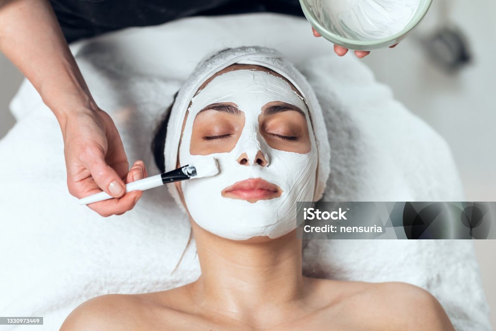 Cosmetologist applying the alginates facial mask to woman while lying on a stretcher in the spa center. Shot of cosmetologist applying the alginates facial mask to woman while lying on a stretcher in the spa center. Facial Mask - Beauty Product Stock Photo