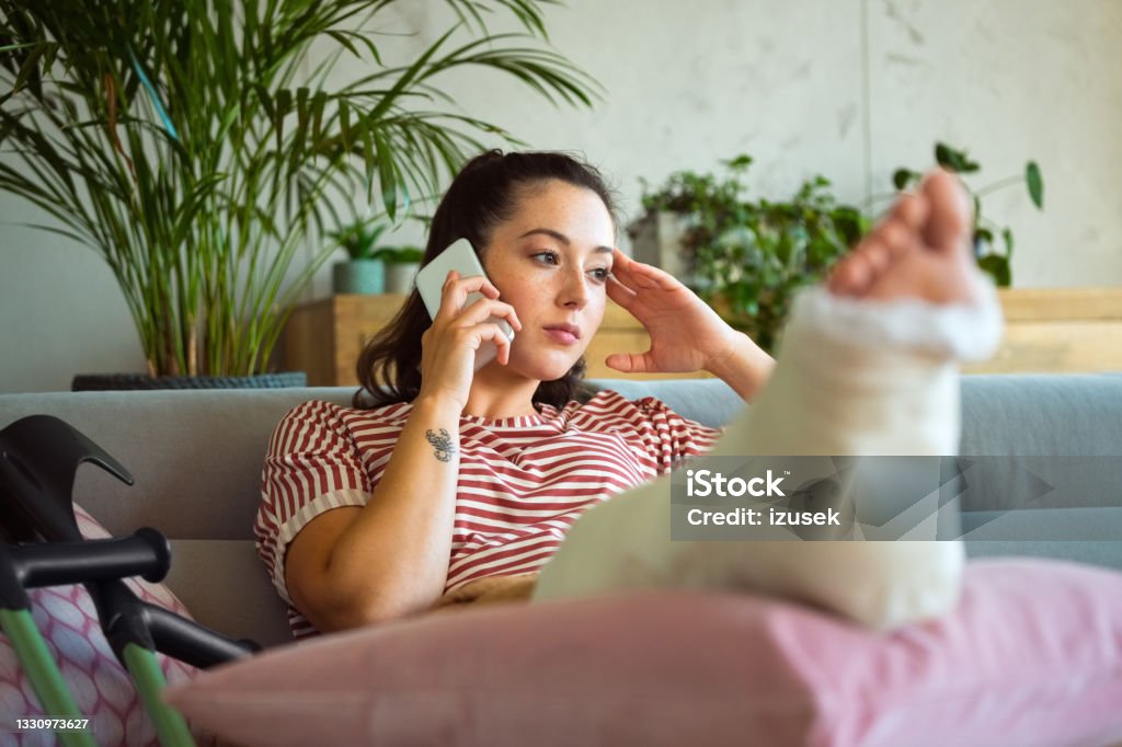 Sad young woman with broken leg on phone Worried young man with broken leg in plaster cast lying down on sofa at home and talking on smart phone. Broken Leg Stock Photo