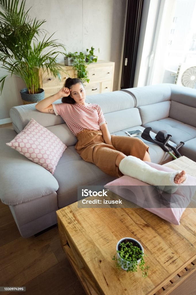 Young woman with broken leg at home Young man with broken leg in plaster cast lying down on sofa at home, smiling at camera. High angle view. Broken Leg Stock Photo