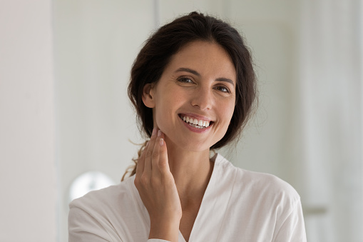Young hispanic female with perfect healthy white smile pose for portrait in bathroom touch neck enjoy morning daily spa procedures. Happy beautiful millennial woman look at camera take care of skin