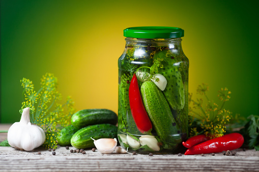 Pickled cucumbers in glass jars and spices and vegetables for preparation of pickles on green background.
