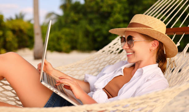 istock Working at sunset. Side view of young happy female using laptop while lying in the hammock on the beach 1330983302
