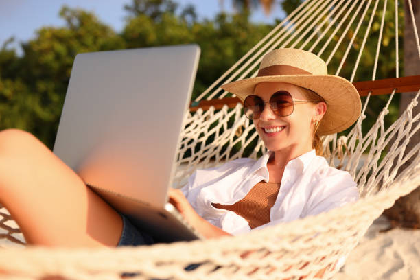 istock Working at sunset. Side view of young happy female using laptop while lying in the hammock on the beach 1330983356