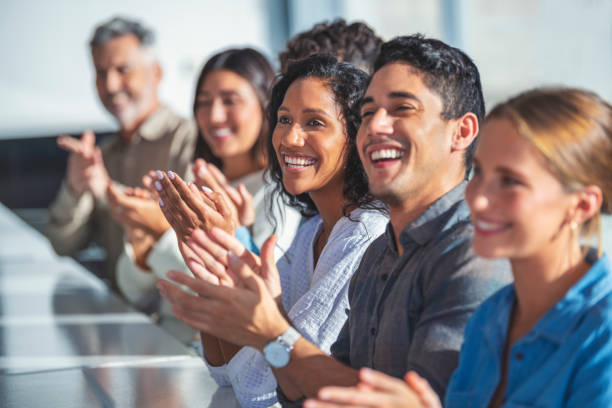 Group of business people applauding a presentation. Group of business people applauding a presentation. They are sitting at a table in a sunny room. There are several ethnicities present including Caucasian, African and Latino employee stock pictures, royalty-free photos & images