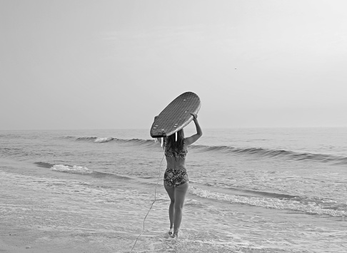 Female surfer with long hair walks toward the ocean with her blue surfboard on her head.