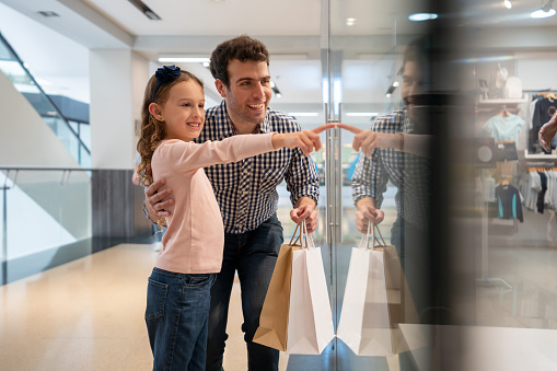 Happy Latin American father shopping at the mall with his daughter and pointing at a shop window - lifestyle concepts