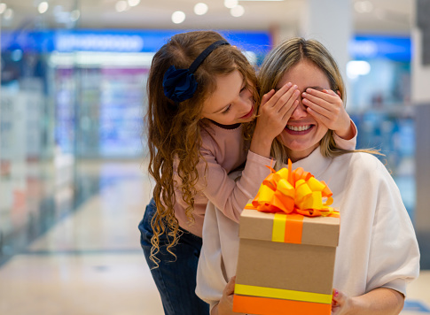 Sweet daughter surprising her mother with a gift at the mall and covering her eyes with her hands - Mother's Day concepts