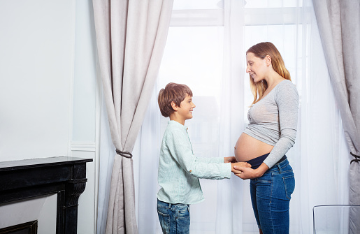 Portrait of a pregnant woman with son holding hands touching tummy near the window