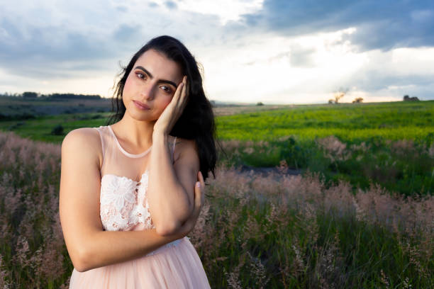 Beautiful Latin woman, wearing a transparent dress of light colors, in the middle of a field of grass, showing tranquility and serenity in a sunset.