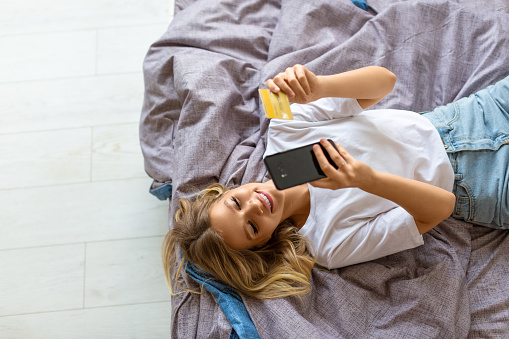 Top vie of a woman lying in bed makes an order through a mobile application. She dials the card number for online purchases. Online shopping concept.