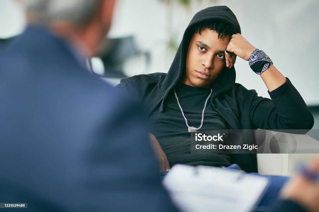 African American teenager having a meeting with psychotherapists at counselling center. Black male adolescent listening to his counsellor during therapy session at psychotherapist's. Rebellion Stock Photo