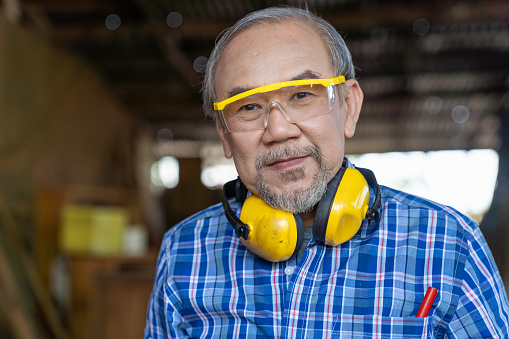 Closeup of Asian senior male carpenter worker wearing safety goggles and earmuffs at work. Asian male woodworker. Asian male craftsman