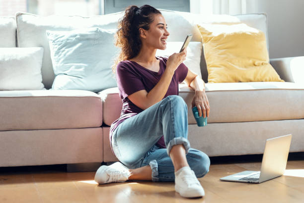 istock Pretty young woman using her mobile phone while working with laptop sitting on the floor at home. 1331037115