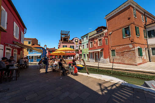 Burano, Italy - June 2th, 2021: Burano island with multi colored houses in front of a small canal with moored boats, restaurants and shops. Venice lagoon, UNESCO world heritage site, Veneto, Italy, Europe. A group of tourists visit the famous island on a sunny spring day with clear sky, while a group of people have lunch in a small outdoor restaurant.