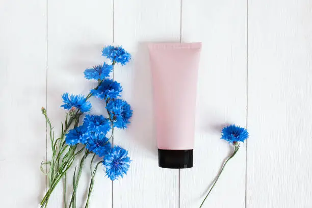 Photo of Top view of mockup pink squeeze bottle plastic tube with black cap and blue cornflowers on a vintage white wooden background. Natural organic spa cosmetics concept. Lovely summer flat lay.