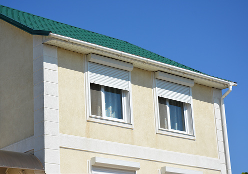 A close-up of a stucco house with a metal green roof, rain gutters, soffit, fascia, and windows with white exterior pvc motorized window roller shutters.
