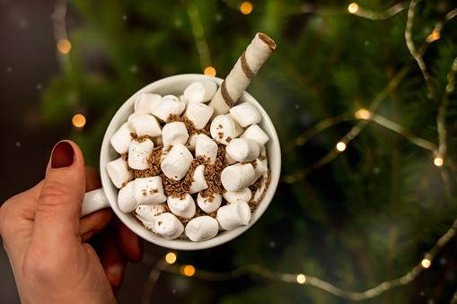 Hot drink with marshmallows and chocolate against a backdrop of blurry Christmas lights.