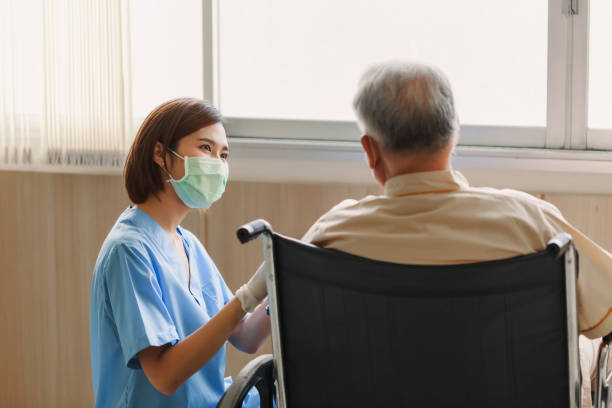 junge asiatische krankenschwester erklärt informationen für älteren mann patient im rollstuhl mit freundlichem smiley im krankenhaus. jugendhilfe mit alten menschen in der altenpflege - asian ethnicity doctor patient healthcare and medicine stock-fotos und bilder